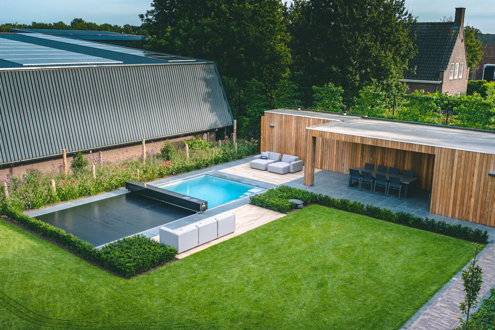 Une piscine entourée de verdure avec une terrasse en bois, des sièges, et un abri adjacent dans un jardin bien entretenu.