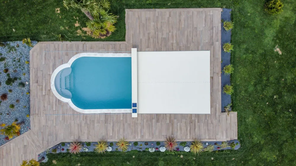Vue aérienne d'une piscine ovale partiellement couverte, entourée de terrasses en bois et de pelouse verte.