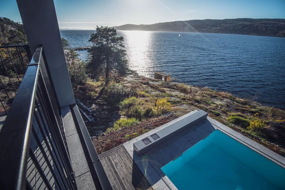 Vue sur une piscine bleue avec terrasse en bois, bordée par un paysage lacustre et une forêt, sous un ciel ensoleillé.
