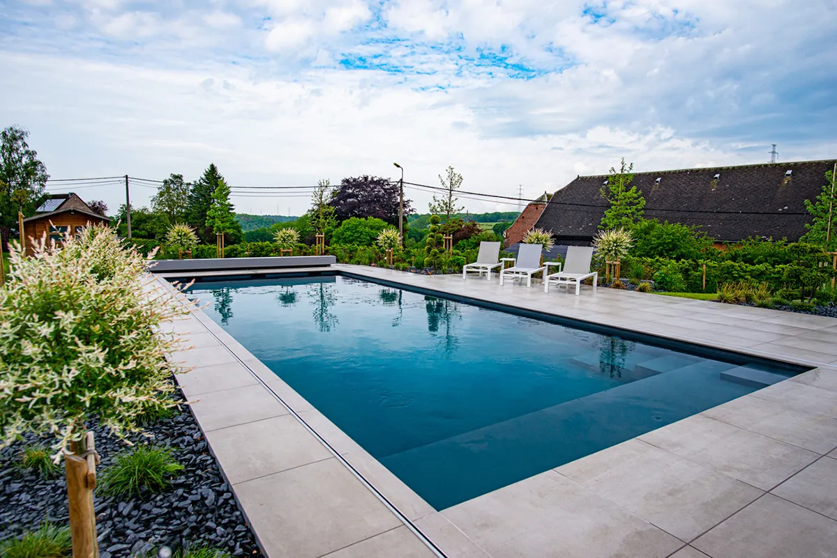 Piscine rectangulaire entourée de dalles grises, avec quatre chaises longues blanches et des arbustes verts en arrière-plan.