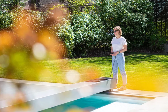 Une personne en t-shirt blanc et jeans utilise une télécommande près d'une piscine entourée de verdure et de feuillage flou.
