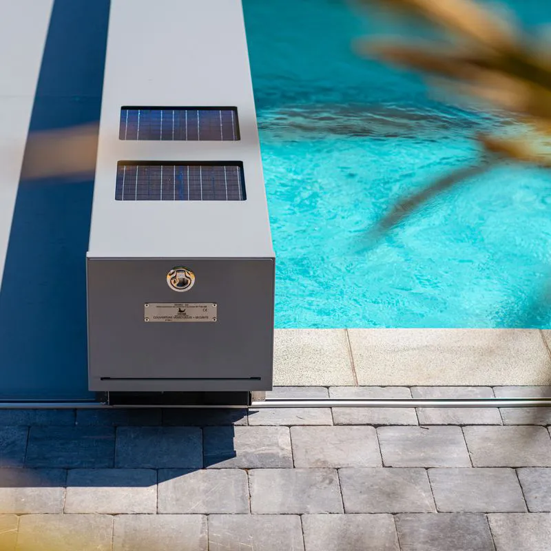 Une couverture de piscine grise Coverseal avec panneaux solaires est installée près d'une piscine aux eaux turquoise, entourée de dalles grises.