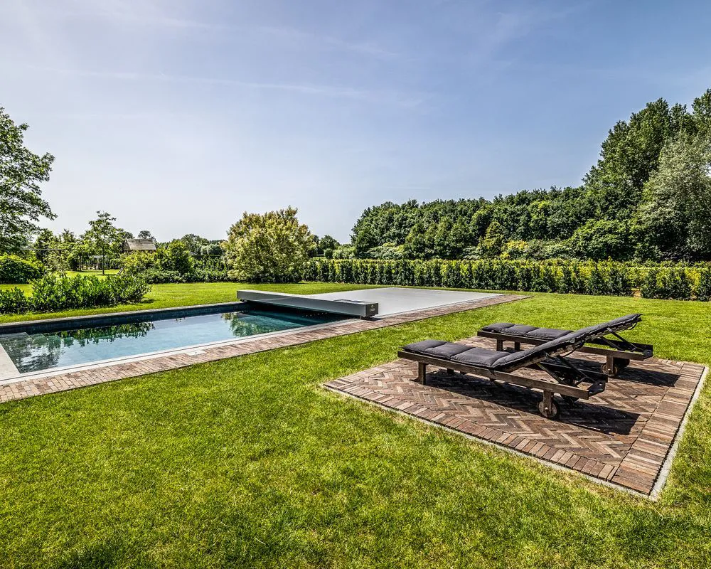 Piscine rectangulaire avec couverture de piscine grise Coverseal, entourée de pelouse verte et de haies, avec deux transats noirs sur une terrasse en bois.