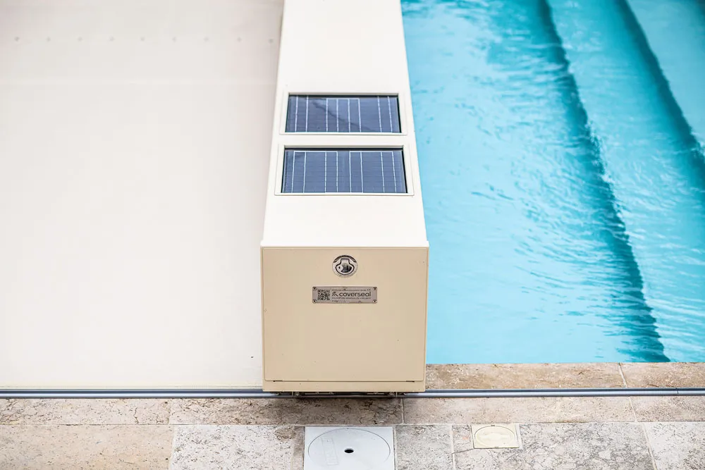 Une couverture de Piscine Coverseal beige avec panneaux solaires se trouve au bord d'une piscine bleue, sur un sol en pierre.