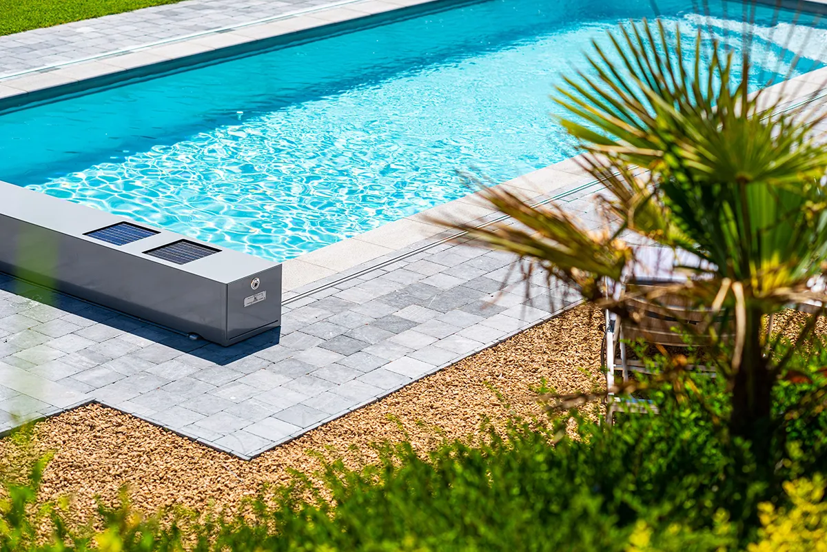 Piscine rectangulaire entourée de dalles grises, galets beige, et palmiers. Dispositif Coverseal gris avec panneaux solaires au bord.