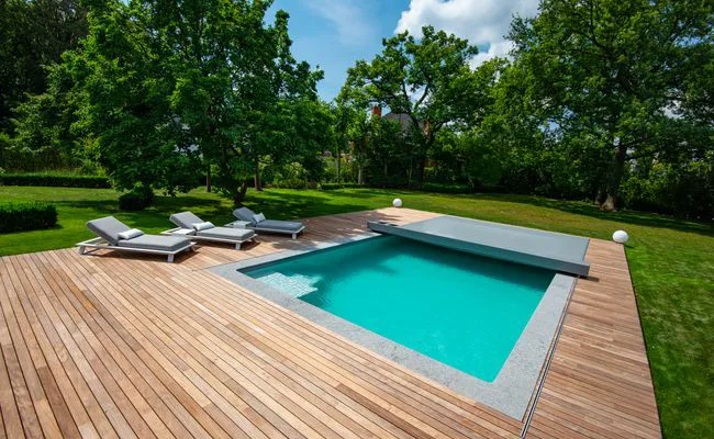 Piscine enterrée entourée d'une terrasse en bois avec trois transats gris, située dans un jardin verdoyant avec des arbres.