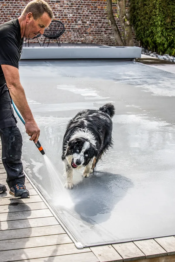 Un homme nettoie une couverture de piscine Coverseal grise avec un tuyau d'arrosage, accompagné d'un chien noir et blanc.