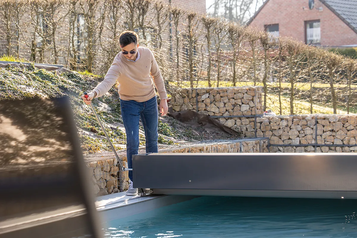 Un homme en pull beige et jeans manipule une perche près d'une piscine et d'une Coverseal entourée de murs en pierre et de haies.