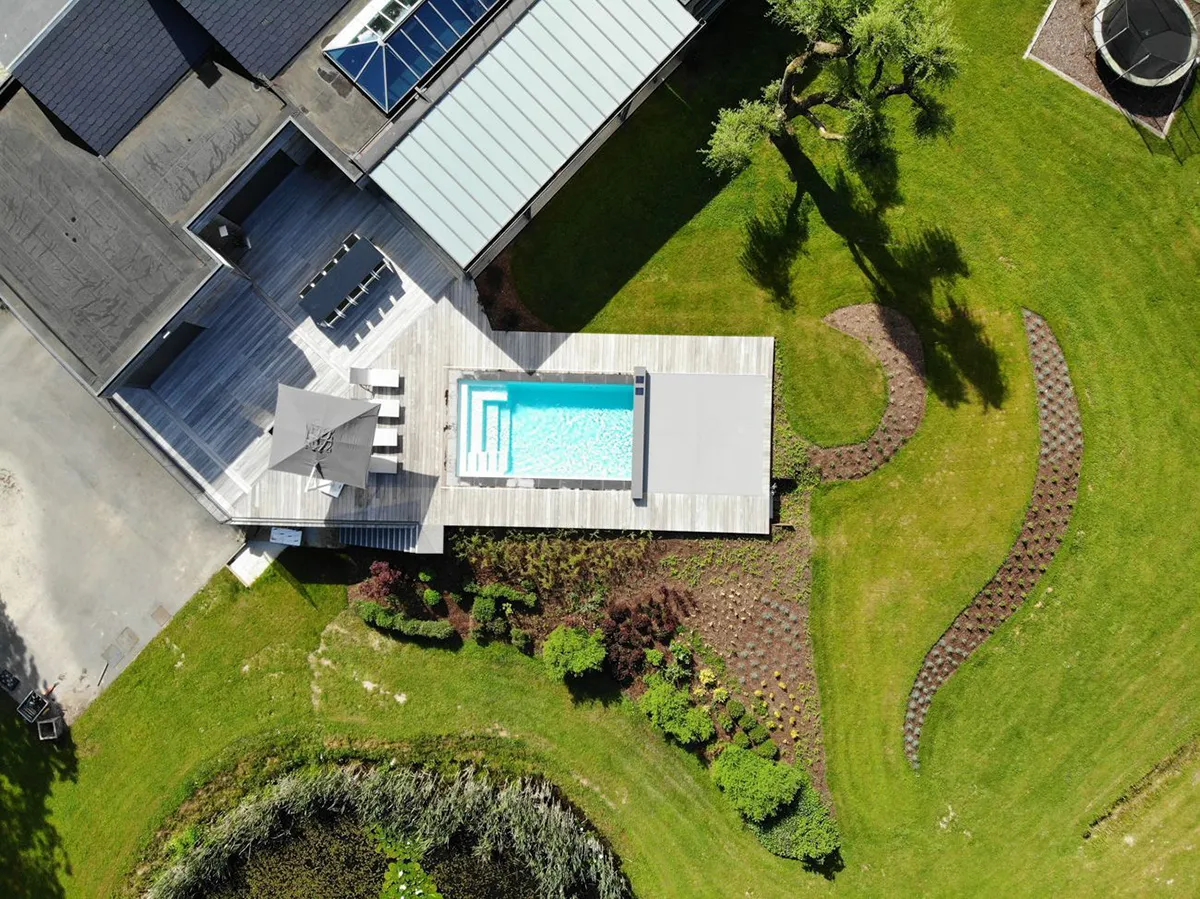 Vue aérienne d'une piscine rectangulaire entourée d'une terrasse en bois grise, avec un jardin verdoyant et un arbre à proximité. La piscine est protégée par une Coverseal