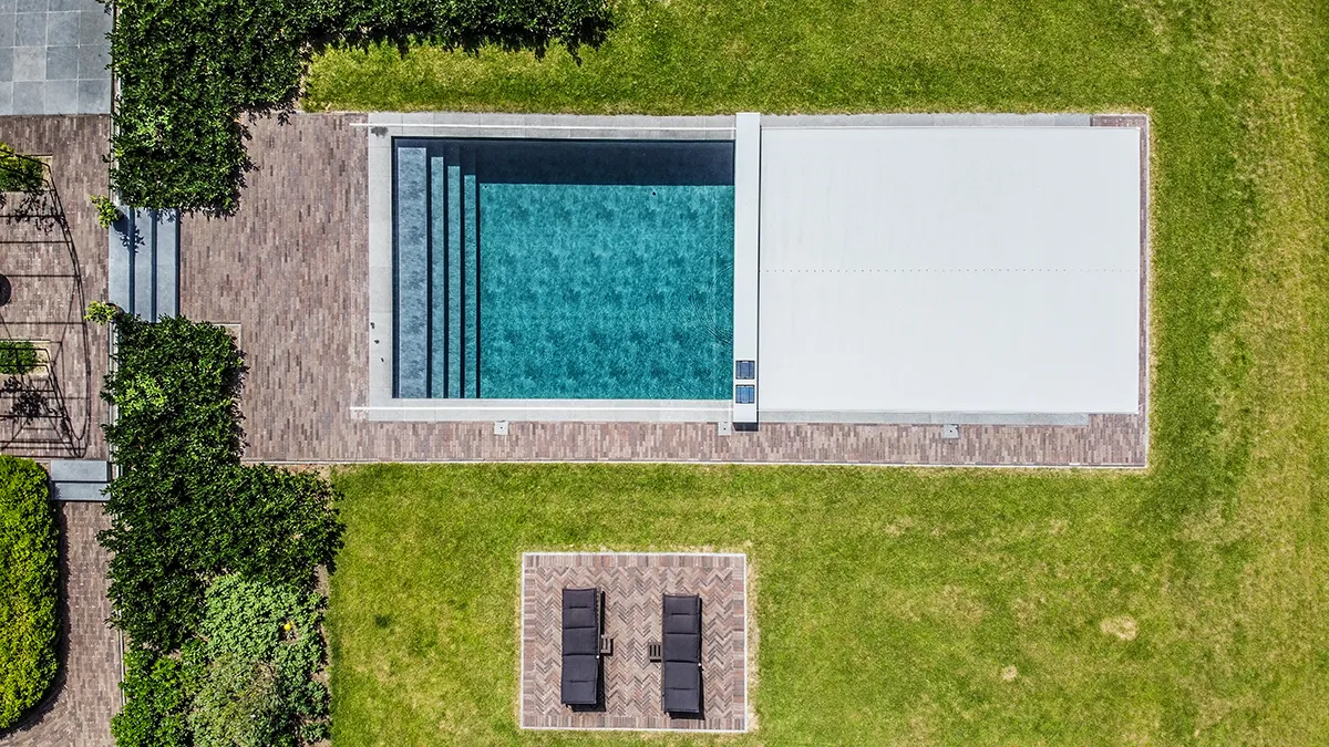 Vue aérienne d'une piscine rectangulaire avec couverture Coverseal, entourée de pavés et de pelouse, et quatre chaises longues noires sur terrasse.