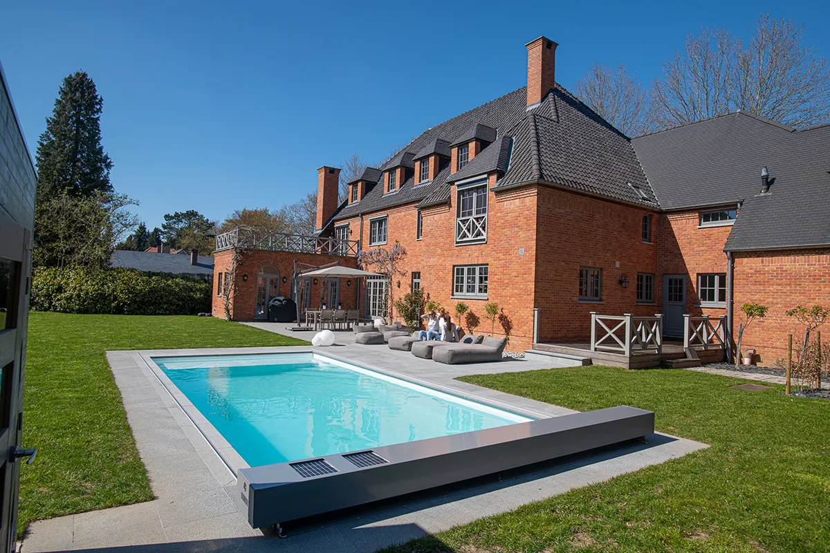 Maison en briques rouges avec toit en ardoise, bordée d'une piscine rectangulaire et d'un jardin verdoyant, sous un ciel bleu.