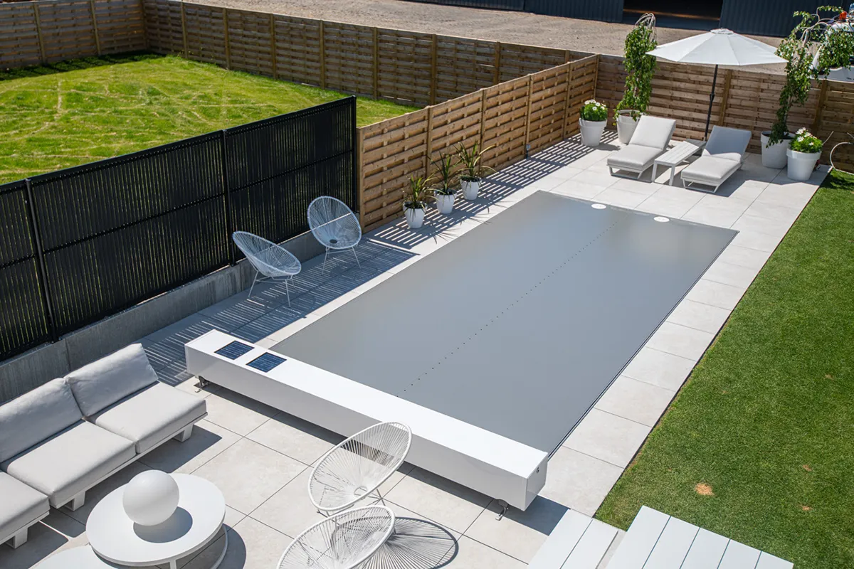 Une piscine couverte d'une Coverseal grise est entourée de chaises longues blanches, de fauteuils et de plantes en pots sur une terrasse carrelée.