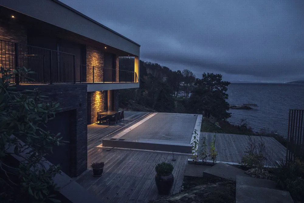 Maison moderne en bord de mer avec terrasse en bois, piscine couverte avec une Coverseal et éclairage extérieur subtil pendant la nuit.