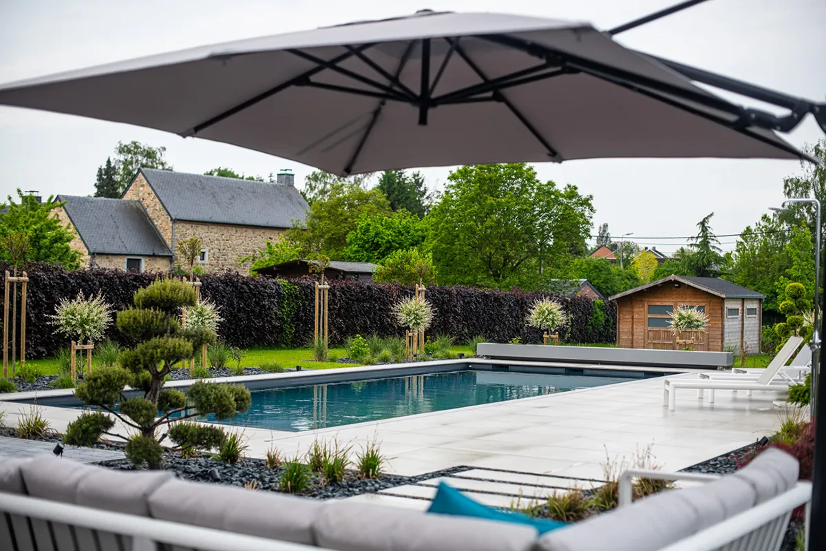 Une piscine entourée de dalles blanches, avec des chaises longues blanches et un grand parasol gris, dans un jardin arboré.