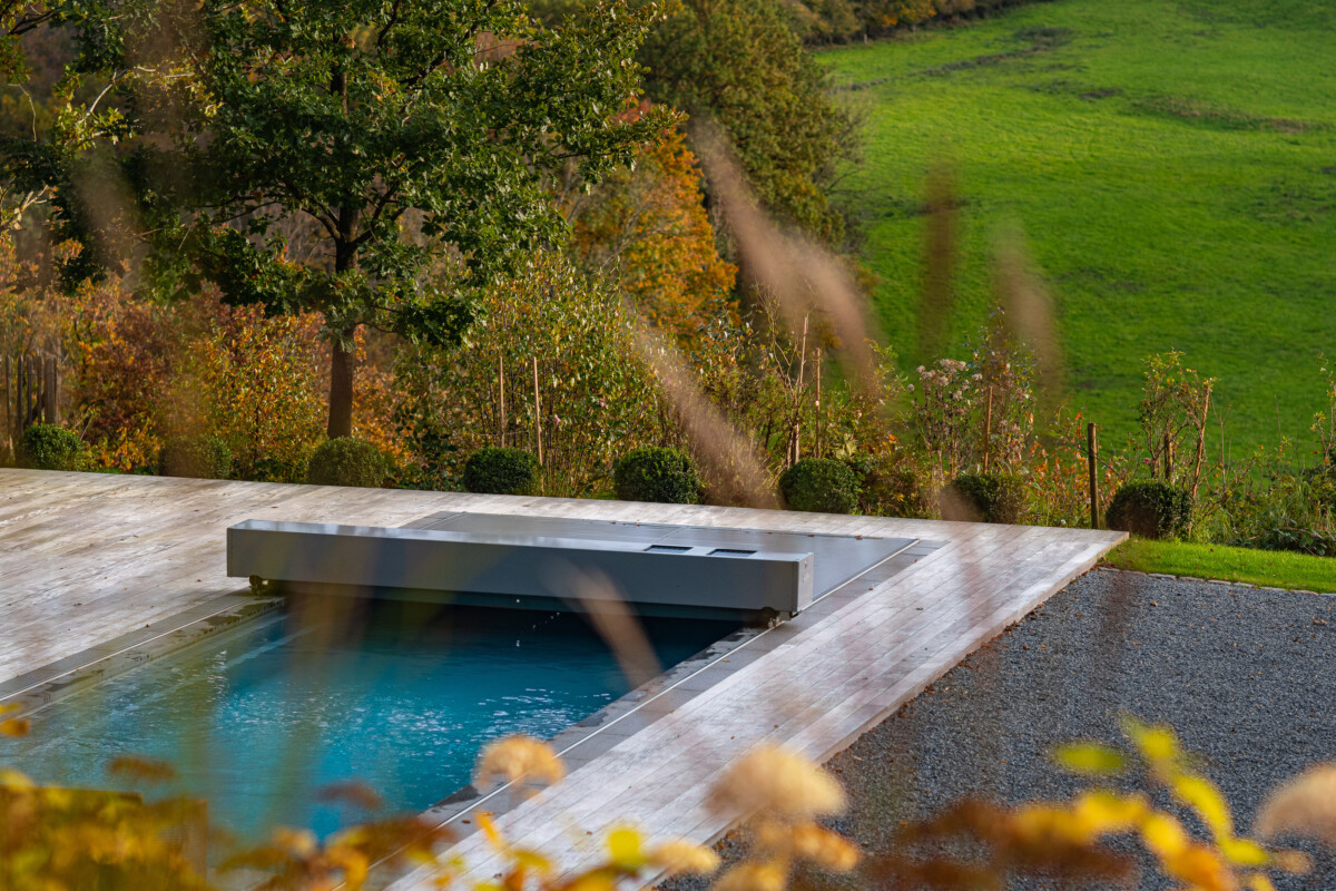 Une piscine couverte rectangulaire est entourée d'une terrasse en bois, avec un arbre et une pelouse en arrière-plan.