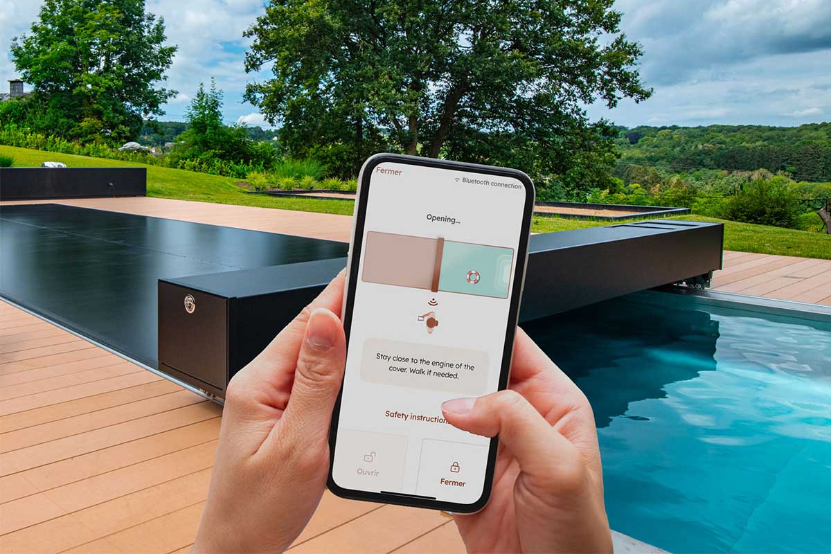 Un smartphone affiche une application contrôlant la couverture d'une piscine, entourée de verdure et d'un ciel nuageux.