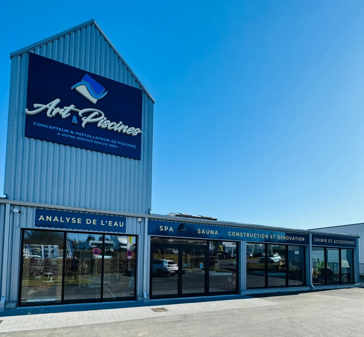 Bâtiment gris avec enseigne "Art et Piscines", vitrines affichant services comme spa, sauna et analyse de l'eau, sous un ciel bleu.