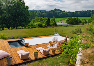 Piscine avec couverture Automatique fermée, terrasse en bois aménagée avec fauteuils et coussins, dans un décor champêtre vallonné.