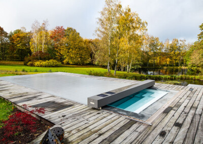 Pond Piscine rectangulaire équipée d’une couverture Coverseal grise, entourée d’une terrasse en bois, avec un grand jardin verdoyant et un étang en arrière-plan.