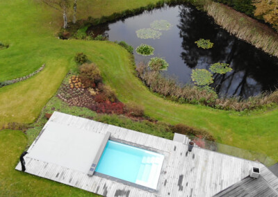 Pond6 Vue aérienne d’une piscine avec couverture Coverseal et terrasse en bois, installée dans un jardin paysager à côté d’un grand étang.