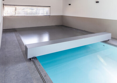 Une couverture de piscine motorisée grise en cours de fermeture sur une piscine intérieure aux lignes modernes.