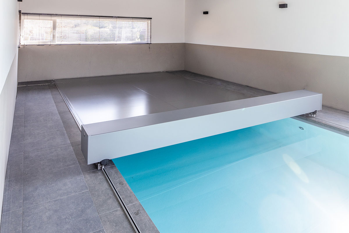 Une couverture de piscine motorisée grise en cours de fermeture sur une piscine intérieure aux lignes modernes.