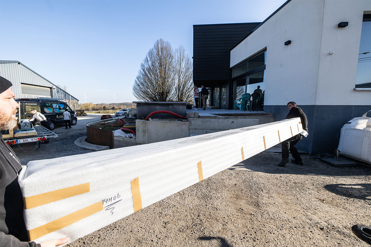 Jade&Noah2 Deux techniciens transportent l'habillage de la Coverseal emballée en mousse blanche devant une maison moderne en construction.