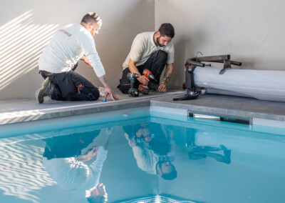 Deux installateurs en tenue de travail posent les rails au bord d’une piscine intérieure, outils électriques à la main.