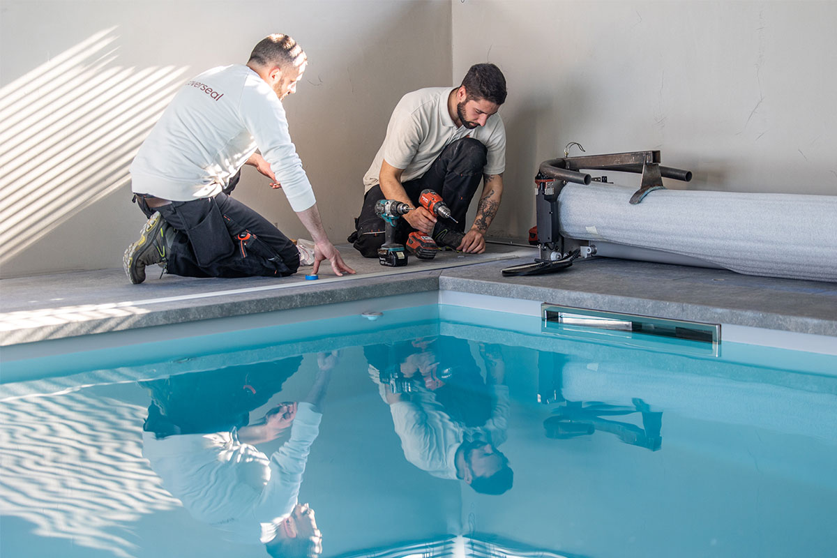 Jade&Noah3 Deux installateurs en tenue de travail posent les rails au bord d’une piscine intérieure, outils électriques à la main.