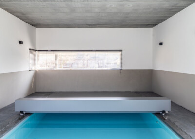 Piscine intérieure couverte par une couverture de piscine gris métal dans une pièce aux murs blancs et sol en carrelage gris.