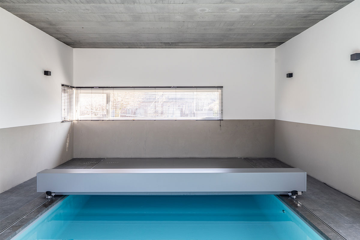 Jade&Noah4 Piscine intérieure couverte par une couverture de piscine gris métal dans une pièce aux murs blancs et sol en carrelage gris.