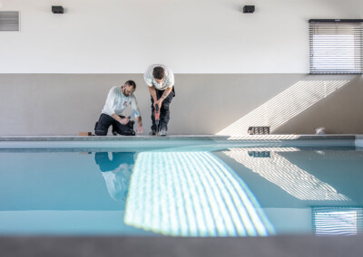 Deux techniciens installent le mécanisme d’une couverture de piscine au bord de l’eau, la lumière du soleil entrant par les stores.