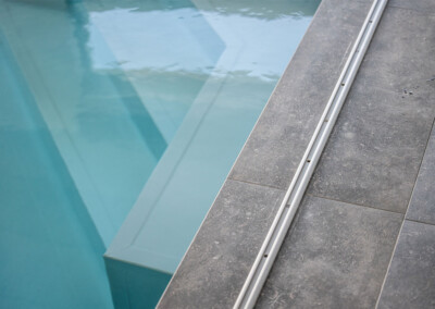 Rail métallique encastré dans le carrelage gris longeant une piscine d’intérieur à l’eau turquoise.