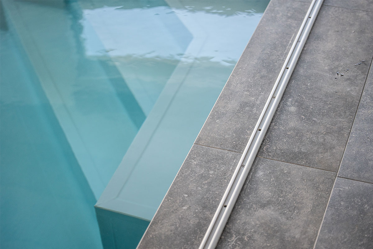Jade&Noah6 Rail métallique encastré dans le carrelage gris longeant une piscine d’intérieur à l’eau turquoise.