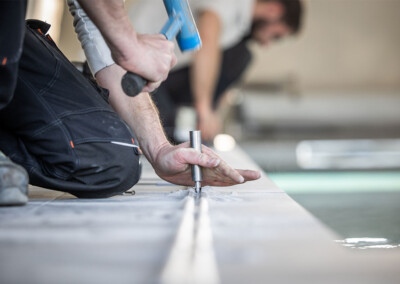 Plan rapproché d’un technicien fixant un rail métallique au sol avec un marteau et un poinçon, lors de l’installation d’une couverture de piscine.