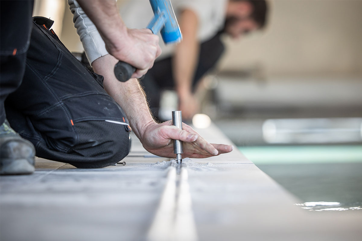 Jade&Noah7 Plan rapproché d’un technicien fixant un rail métallique au sol avec un marteau et un poinçon, lors de l’installation d’une couverture de piscine.